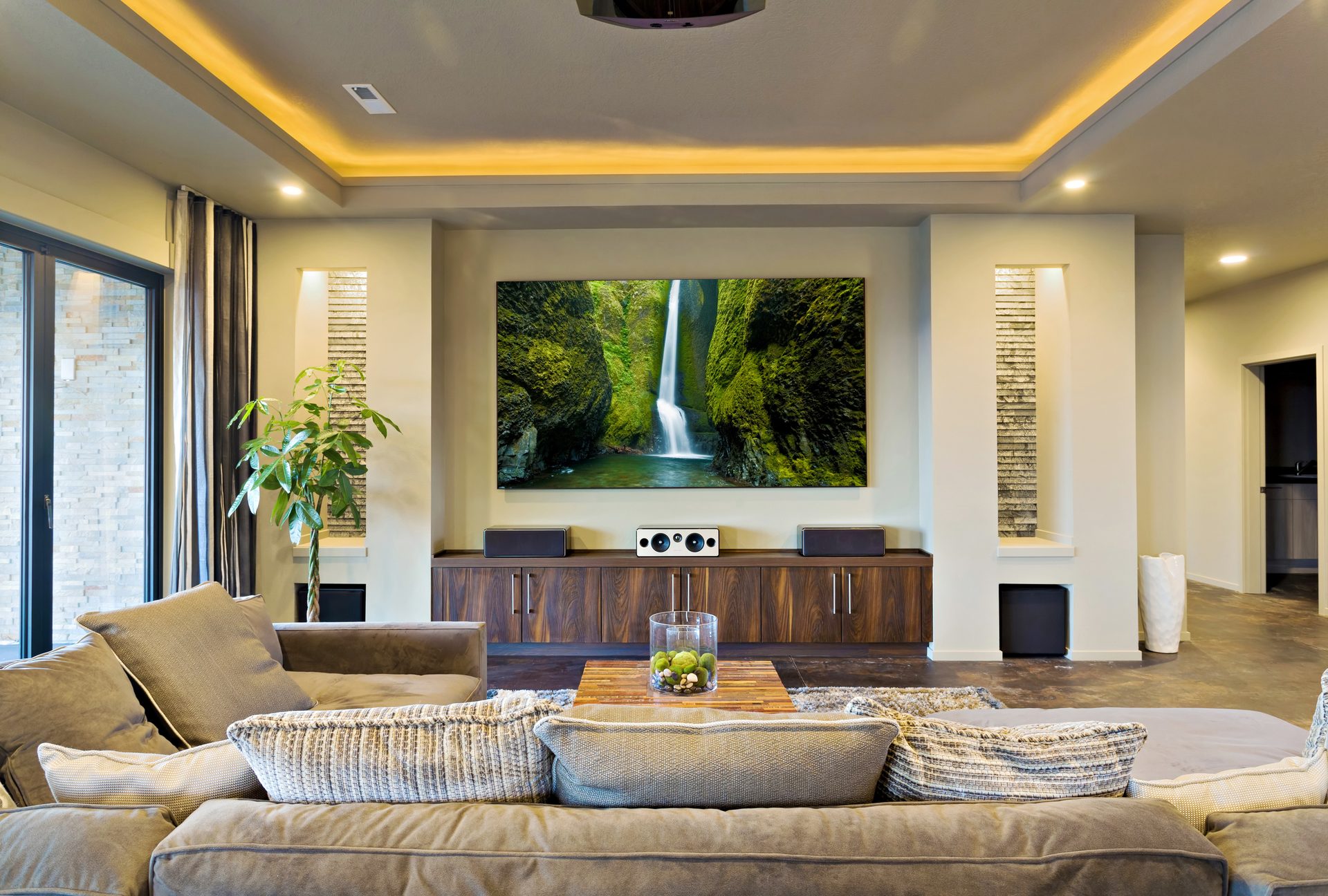 A lounge with warm LED strip lighting hidden in a recessed ceiling tray, downlights, and a large wall-mounted screen above a timber cabinet with speakers